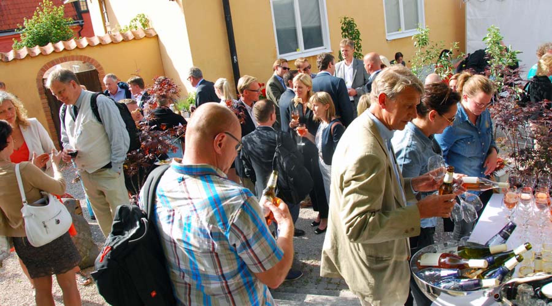 Ett mingelevent i solen. Människor tar vin från en buffé, står i smågrupper och pratar med varandra.